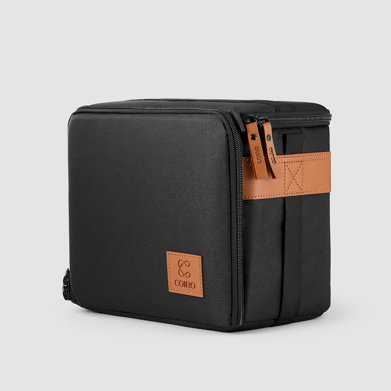 May include: Black rectangular bag with a brown leather strap and zip. The bag has a small brown square patch with the word "COIRO" printed on it. The bag is designed to carry items.
