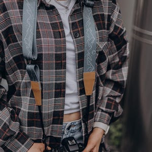 May include: A gray camera strap with a white mountain design and brown leather accents is draped over a plaid shirt. The camera strap is attached to a black Canon camera. The shirt is a mix of black, brown, and red.
