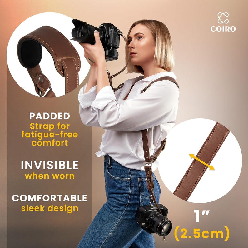 May include: Brown leather camera strap with a padded shoulder section. The strap is 1 inch (2.5 cm) wide and has a sleek design. The strap is worn by a person holding a camera.