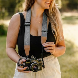 May include: A black Nikon film camera with a gray and brown camera strap featuring a mountain design. The camera strap is around the neck. The camera has a lens and is held by a person.