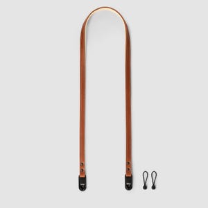 Puede incluir: Una correa de cámara de cuero marrón con herrajes de metal negro y dos clips negros. La correa está hecha de cuero liso cosido y está diseñada para usarse alrededor del cuello o el hombro.