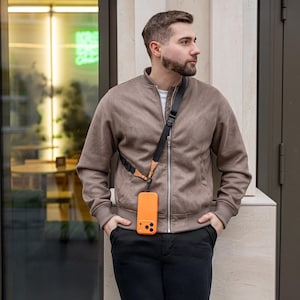 May include: An orange phone case with a black strap is worn across the body. The case is attached to a black and brown strap. The person is wearing a brown bomber jacket and black pants.