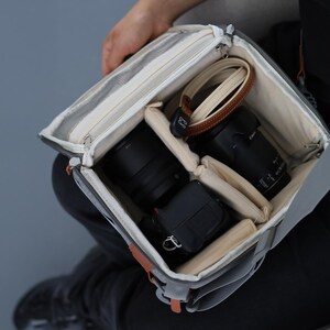May include: A grey camera bag with a beige interior, containing a camera, lenses, and a brown and beige camera strap. The bag has a zip closure and orange leather accents.
