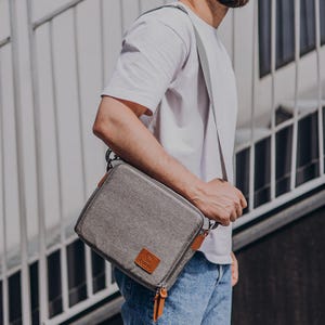 May include: A grey messenger bag with brown leather detailing and a zip pull. The bag is being carried over the shoulder by a person wearing a white t-shirt and blue jeans.