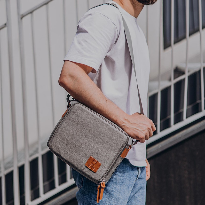 May include: A grey messenger bag with brown leather detailing and a zip pull. The bag is being carried over the shoulder by a person wearing a white t-shirt and blue jeans.