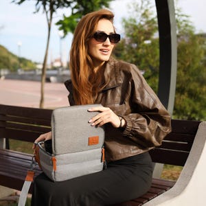 May include: A grey travel bag with brown leather detailing and orange zip pulls. The bag has a shoulder strap and a rectangular shape. The bag is open, revealing the interior. The bag has a brown leather patch with a logo.