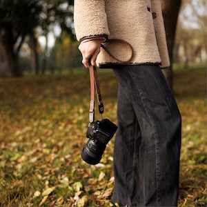 May include: A brown leather camera strap with a black camera hanging from it. The strap is held by a person wearing a tan jacket and dark jeans. The background is a blurred outdoor scene with fallen leaves.