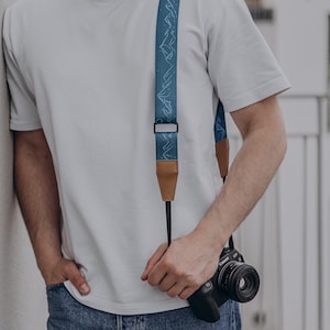 May include: A teal camera strap with a white mountain range pattern. The strap has brown leather ends and a black connector. A black camera is attached to the strap.