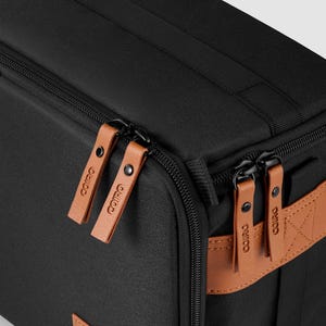 May include: Black rectangular bag with brown leather detailing. It has brown leather zipper pulls and a brown leather patch with the brand logo. The bag features a black handle and a black zip.
