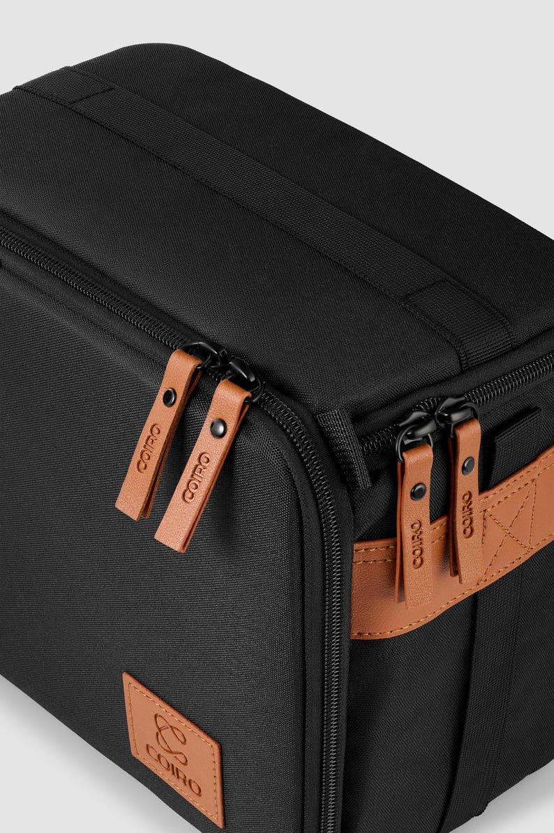 May include: Black rectangular bag with brown leather detailing. It has brown leather zipper pulls and a brown leather patch with the brand logo. The bag features a black handle and a black zip.