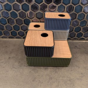 May include: A stack of four tissue box holders in various colors. The boxes have a rectangular shape with a wooden top and a ribbed, colored base. Colors include green, black, gray, and blue. The top has a circular opening for tissue dispensing.