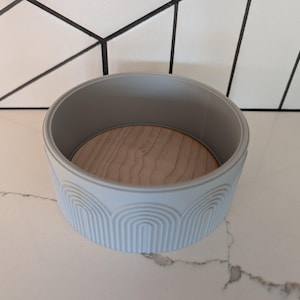 May include: A light blue, round container with a wooden base. The container has a ribbed, arched design around the exterior. The interior is a light gray color. The container is sitting on a white marble surface.