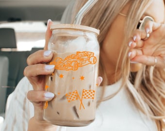 May include: A clear glass tumbler with a bamboo lid and a clear straw, filled with a beverage. The tumbler features an orange outline design of a race car, stars, and checkered flags. The drink is held by a person.