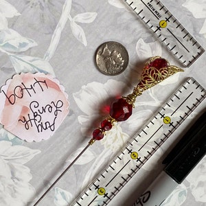 May include: A red and gold beaded hat pin with a decorative filigree top. The pin is about 4 inches long and is shown next to a ruler and a black marker.