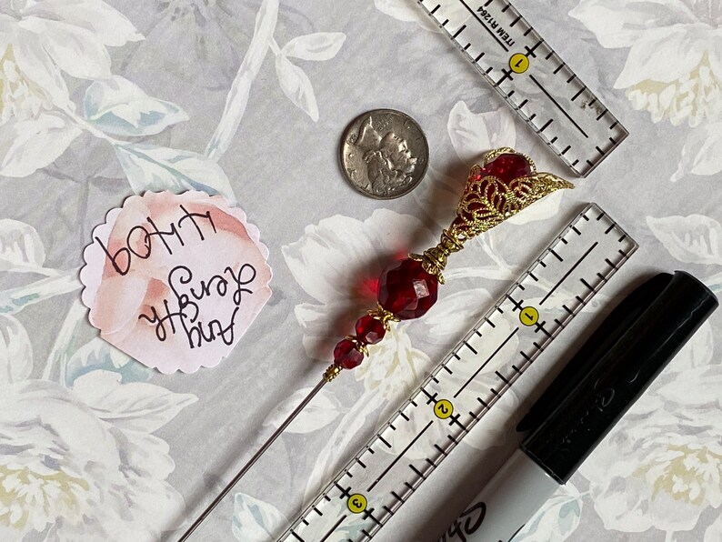 May include: A red and gold beaded hat pin with a decorative filigree top. The pin is about 4 inches long and is shown next to a ruler and a black marker.