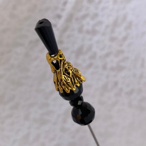 May include: A black and gold decorative pin with a black crystal top and a gold filigree design. The pin is on a white background.