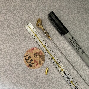 以下が含まれることがあります： ラインストーンが付いた金色の花のピン、金色の金属製コネクター、長く細い銀色のピン。ピンは、「Omnigrid」と「USA」の文字が印刷された定規で測定されています。紙に「1336」という数字が見えます。