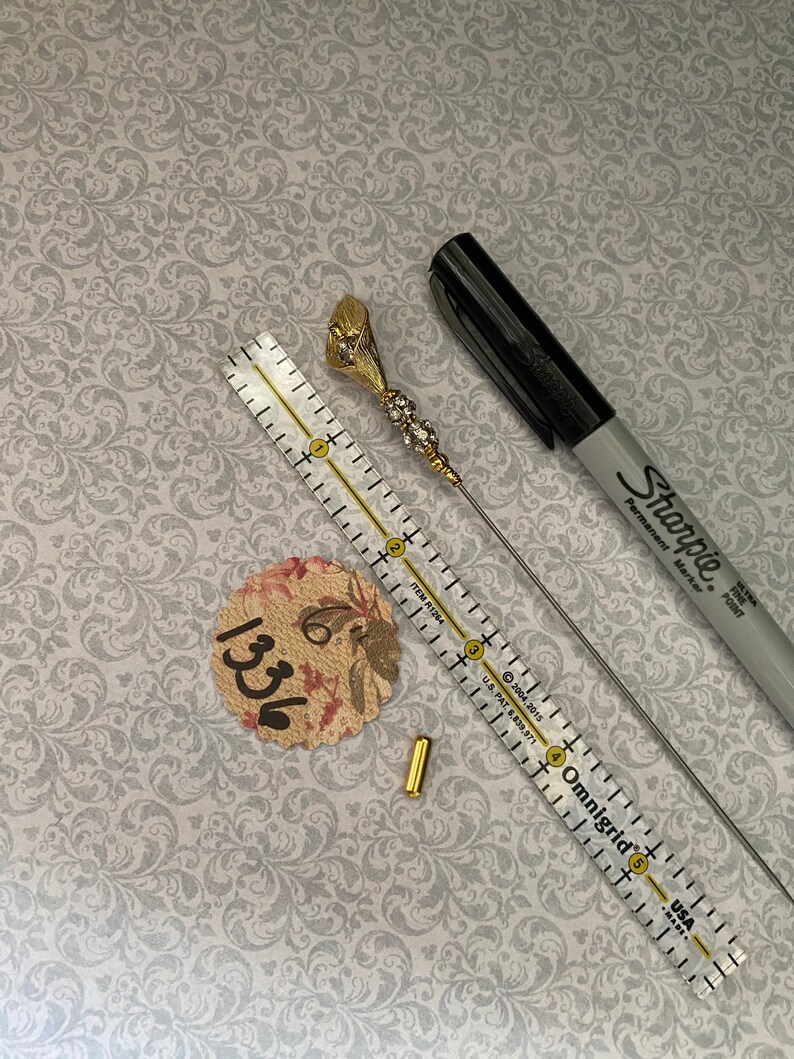 以下が含まれることがあります： ラインストーンが付いた金色の花のピン、金色の金属製コネクター、長く細い銀色のピン。ピンは、「Omnigrid」と「USA」の文字が印刷された定規で測定されています。紙に「1336」という数字が見えます。