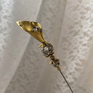 May include: A gold-toned metal stick pin featuring a calla lily flower head with a textured finish. The stem is adorned with two spherical beads encrusted with clear, sparkling rhinestones, separated by small gold-toned metal floral accents.