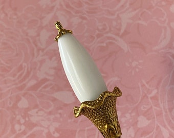 May include: A white ceramic pin with a gold metal base and a decorative floral design. The pin has a long, thin stem.