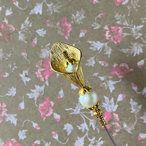 May include: Gold-toned metal pin with a white pearl and a stylized flower design. The flower is shaped like a calla lily.