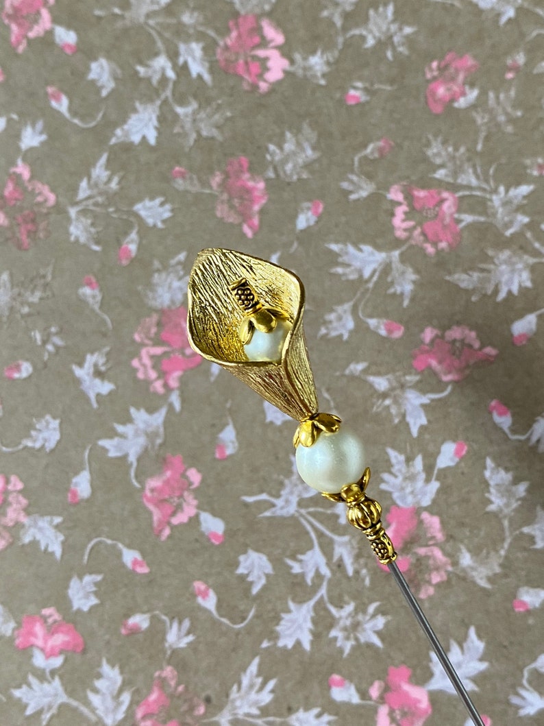 May include: Gold-toned metal pin with a white pearl and a stylized flower design. The flower is shaped like a calla lily.