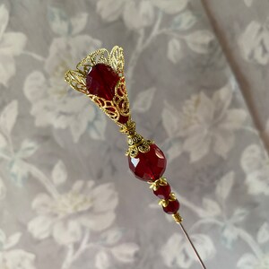 May include: A gold and red decorative pin with a filigree design and a long, thin needle. The pin features a large, round red bead and a smaller red bead with gold accents.