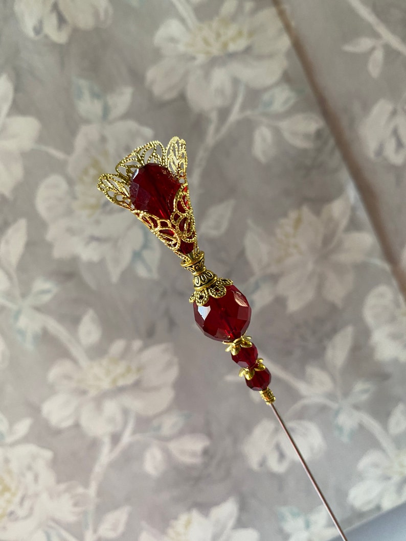May include: A gold and red decorative pin with a filigree design and a long, thin needle. The pin features a large, round red bead and a smaller red bead with gold accents.