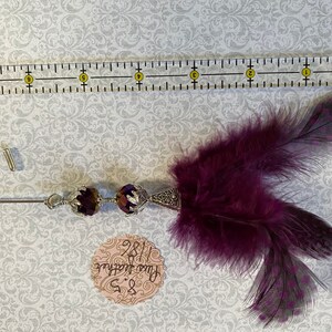 K&ouml;nnte beinhalten: Dekorative Anstecknadel mit einem dunkelvioletten Federakzent. Die Nadel hat zwei dunkelviolette facettierte Perlen und ein silberfarbenes Metalldesign. Ein Lineal liegt neben der Nadel, und ein kleines Etikett mit Text ist ebenfalls sichtbar.