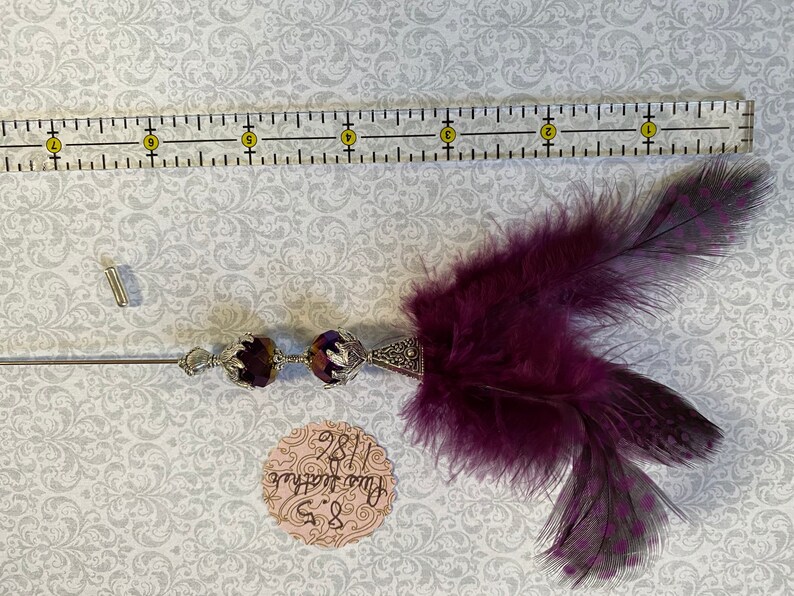 K&ouml;nnte beinhalten: Dekorative Anstecknadel mit einem dunkelvioletten Federakzent. Die Nadel hat zwei dunkelviolette facettierte Perlen und ein silberfarbenes Metalldesign. Ein Lineal liegt neben der Nadel, und ein kleines Etikett mit Text ist ebenfalls sichtbar.