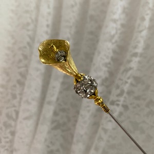 Puede incluir: Un broche floral dorado con dise&ntilde;o de cala. El broche presenta un grupo de cristales transparentes y un fino alfiler plateado. La cala tiene detalles texturizados y el alfiler tiene detalles dorados decorativos.