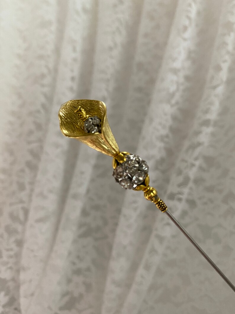 Puede incluir: Un broche floral dorado con dise&ntilde;o de cala. El broche presenta un grupo de cristales transparentes y un fino alfiler plateado. La cala tiene detalles texturizados y el alfiler tiene detalles dorados decorativos.