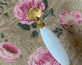 May include: A white and gold decorative pin with a flower design. The pin has a white ceramic base and a gold metal flower with a leaf design.