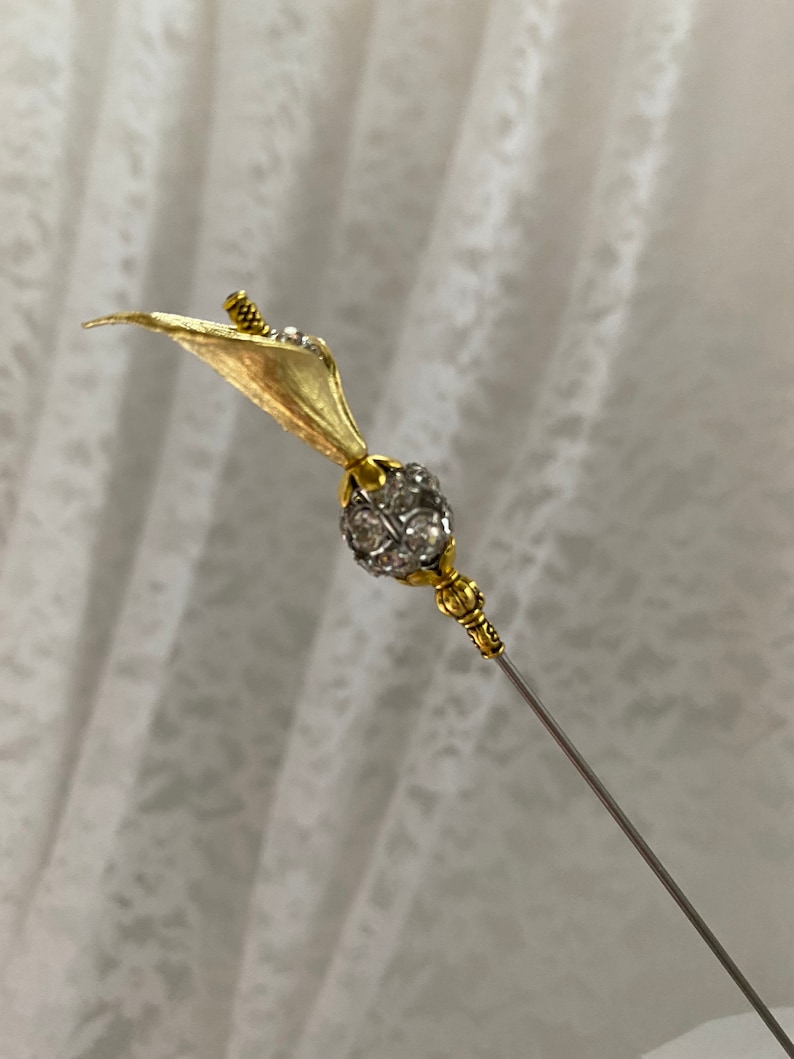Puede incluir: Alfiler de sombrero dorado con dise&ntilde;o de cala. El lirio tiene un p&eacute;talo texturizado y un peque&ntilde;o acento de cristal brillante. El alfiler presenta una bola redonda con incrustaciones de cristal y una aguja larga plateada.
