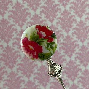 May include: A silver pin with a white round bead featuring a pink floral design. The bead is attached to a silver decorative piece and a silver pin.