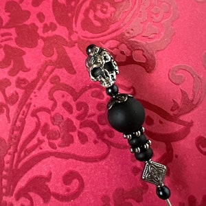 May include: A black and silver skull pin with a black bead and a silver charm. The pin is on a red background.