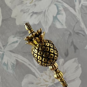 Op de afbeelding: Een gouden decoratieve pin in de vorm van een ananas met een getextureerd oppervlak en een parelbezet steeltje.