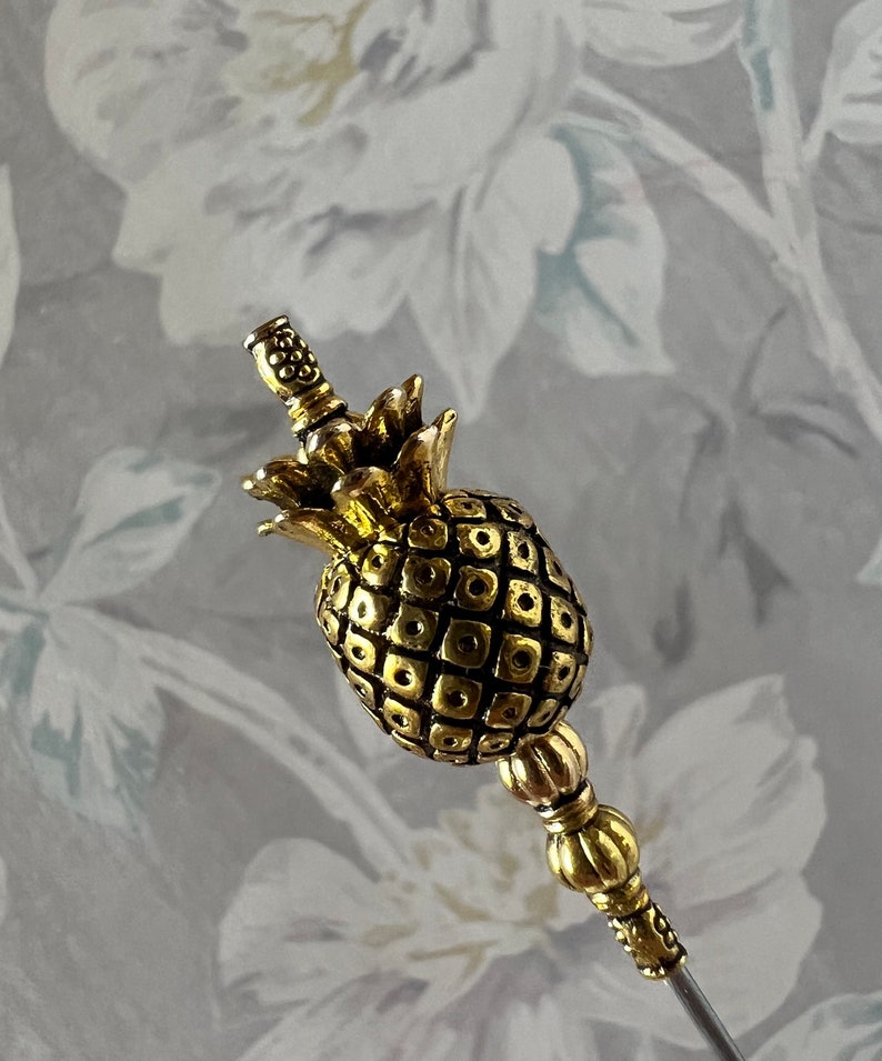 Op de afbeelding: Een gouden decoratieve pin in de vorm van een ananas met een getextureerd oppervlak en een parelbezet steeltje.