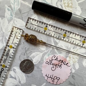 Op de afbeelding: Een gouden ananasvormige broche met een lange, dunne, zilveren steel. De broche wordt gemeten met een liniaal en een munt. De tekst "Choose length 4489" staat op een roze papiertje.
