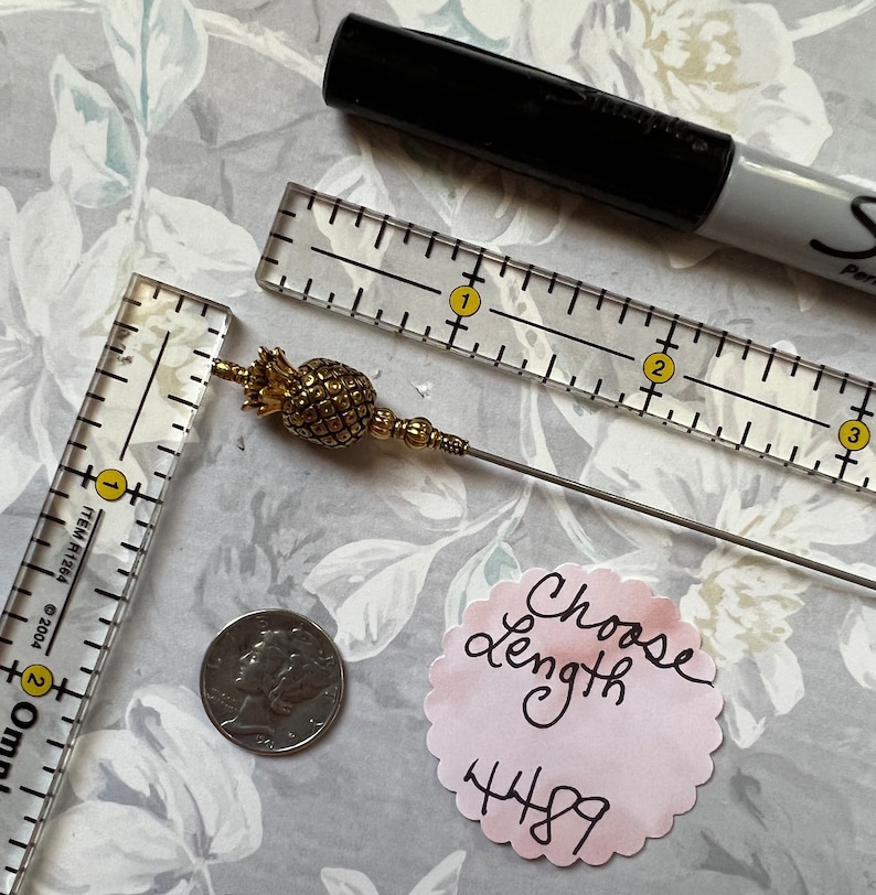 Op de afbeelding: Een gouden ananasvormige broche met een lange, dunne, zilveren steel. De broche wordt gemeten met een liniaal en een munt. De tekst "Choose length 4489" staat op een roze papiertje.