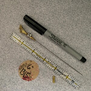 Puede incluir: Un surtido de materiales de manualidades que incluye un rotulador Sharpie negro, un alfiler dorado con una parte superior decorativa, una regla transparente, un peque&ntilde;o tubo dorado y un c&iacute;rculo con estampado floral con el texto manuscrito "6" 1315".