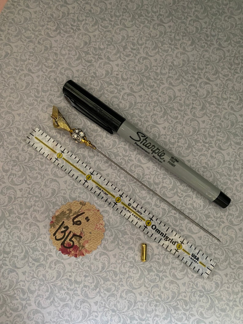 Puede incluir: Un surtido de materiales de manualidades que incluye un rotulador Sharpie negro, un alfiler dorado con una parte superior decorativa, una regla transparente, un peque&ntilde;o tubo dorado y un c&iacute;rculo con estampado floral con el texto manuscrito "6" 1315".