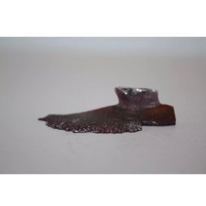 May include: A small, irregularly shaped piece of metal with a dark purple and silver finish. The metal has a rough, textured surface.