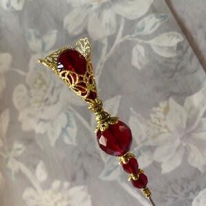 May include: A red glass and gold metal decorative pin with a teardrop-shaped top and a round bead in the middle.
