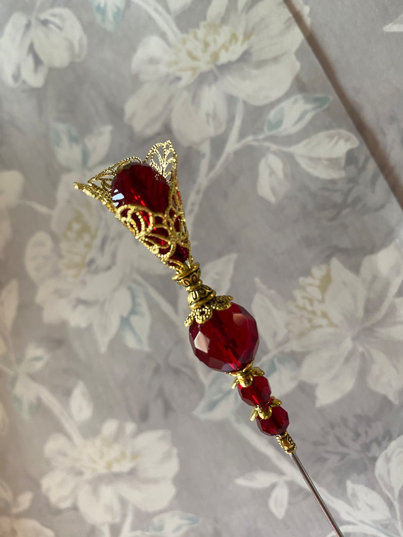 May include: A red glass and gold metal decorative pin with a teardrop-shaped top and a round bead in the middle.
