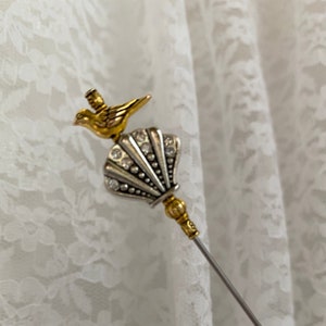 May include: A decorative hat pin featuring a gold-coloured bird atop a silver-coloured shell design, adorned with clear rhinestones. The pin has a long, slender, silver-coloured shaft and gold-coloured accents.