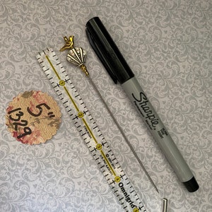 May include: A decorative hat pin with a gold bird and seashell design, a black Sharpie marker, a 12.7 cm ruler, and a floral tag. The pin has a silver needle. The Sharpie is gray and black.