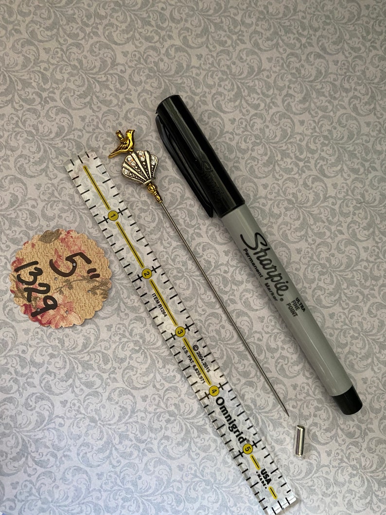 May include: A decorative hat pin with a gold bird and seashell design, a black Sharpie marker, a 12.7 cm ruler, and a floral tag. The pin has a silver needle. The Sharpie is gray and black.