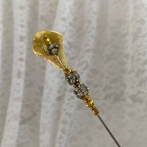 以下が含まれることがあります： クリアなラインストーンのアクセントが付いたゴールドトーンの花のピン。ピンはカラーリリーの形状で、長く細い茎があります。