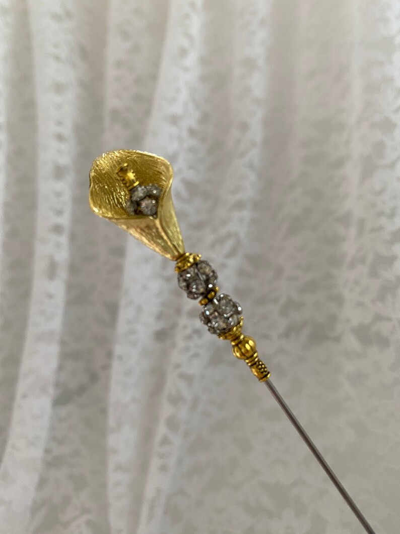以下が含まれることがあります： クリアなラインストーンのアクセントが付いたゴールドトーンの花のピン。ピンはカラーリリーの形状で、長く細い茎があります。
